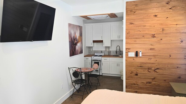 A compact kitchen with white cabinets, a stove, and a sink. There's a small table with chairs and a painting on the wall.