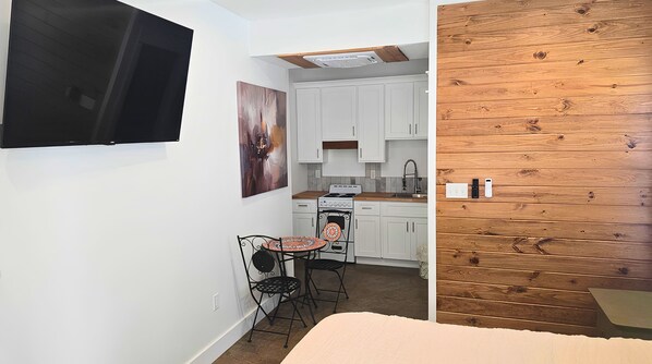 A compact kitchen with white cabinets, a stove, and a sink. There's a small table with chairs and a painting on the wall.