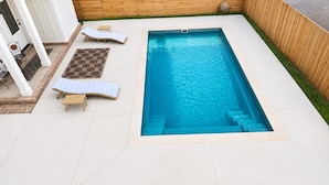 A rectangular swimming pool with a built-in hot tub, surrounded by a white tiled deck, a wooden fence, and a small garden area with a bench and a patterned rug.