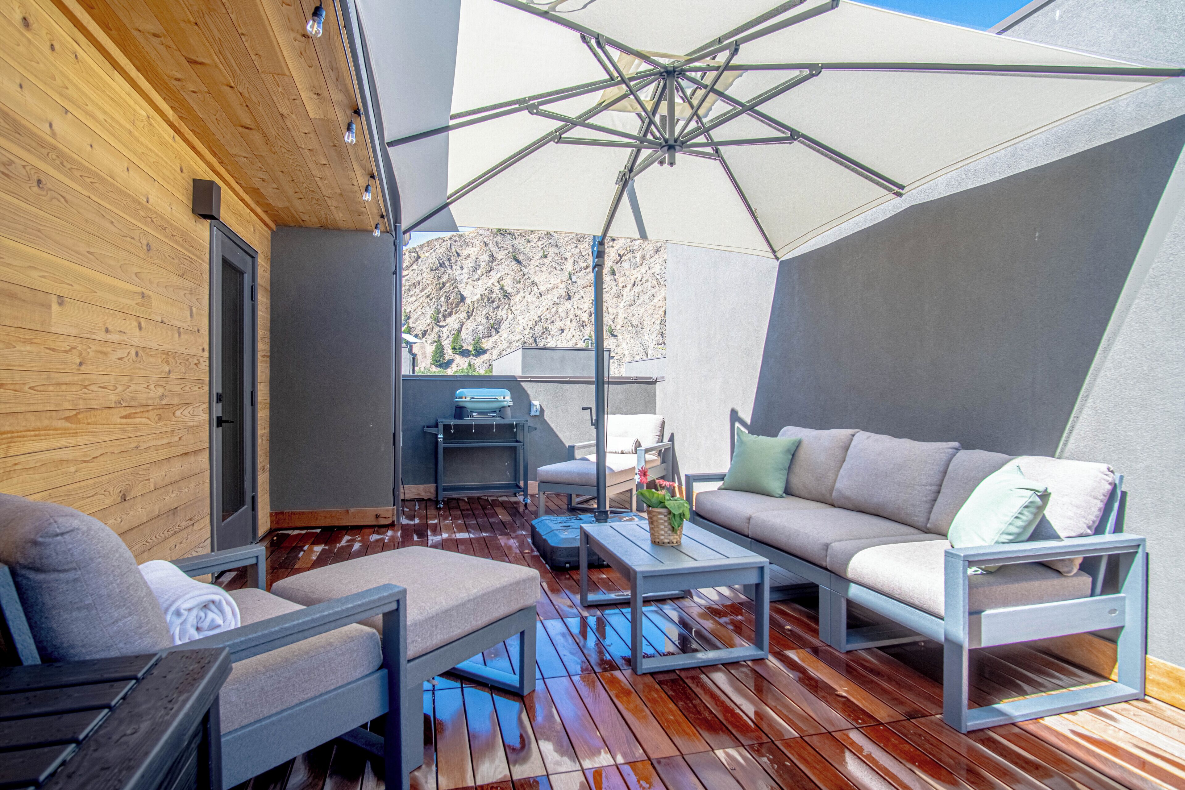 Ein moderner Außenbereich zum Sitzen mit einer Holzterrasse, einem großen Sonnenschirm und Blick auf eine gebirgige Landschaft.
