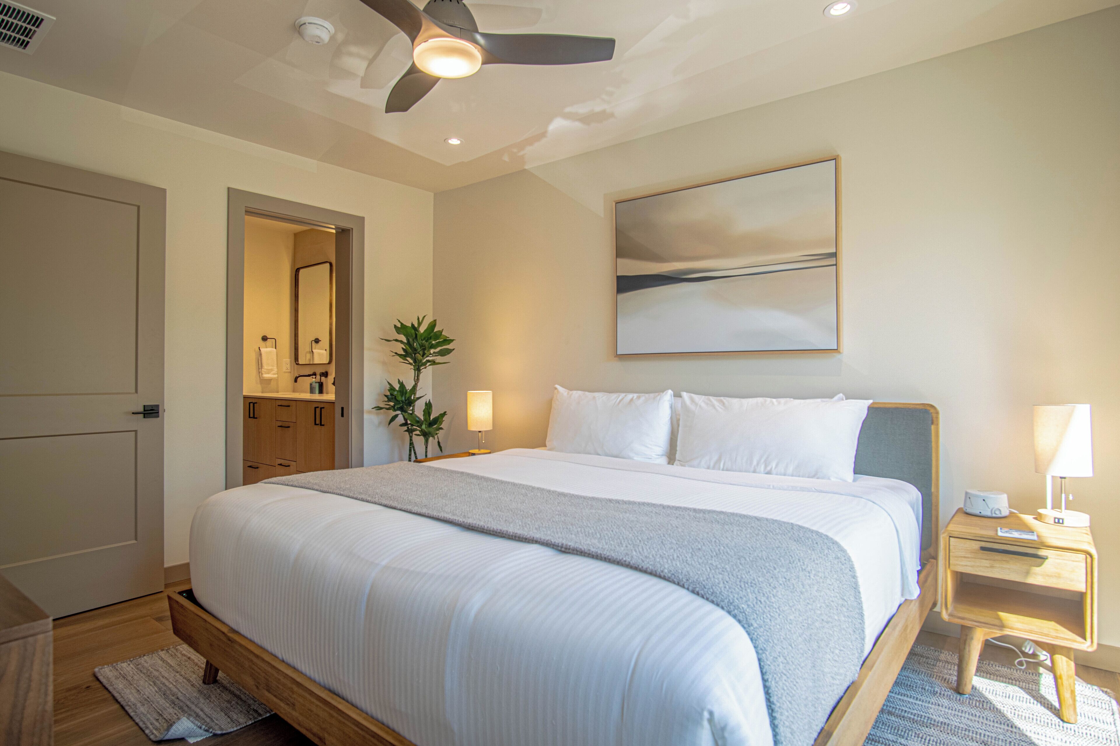 A hotel room with a bed, bedside tables, a ceiling fan, a painting, and a bathroom visible through an open door.