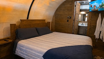 Una habitación con cama, cabecero de madera, mesita de noche, televisión y baño con espejo y lavamanos.