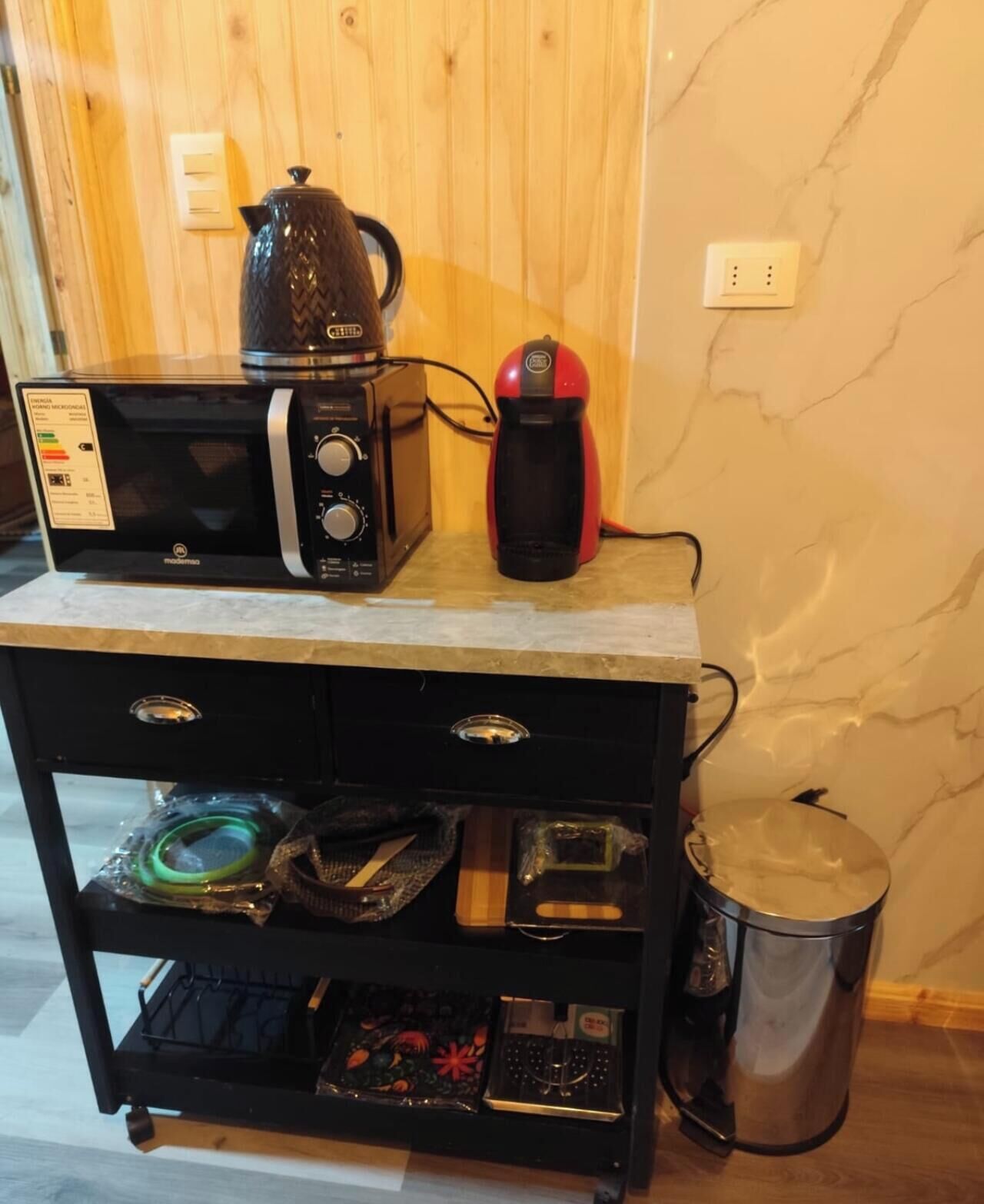 Una encimera de cocina con microondas, cafetera roja y hervidor de acero inoxidable. Debajo de la encimera, hay un mueble negro con cajones y estantes que contienen utensilios de cocina.