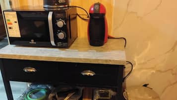 Una encimera de cocina con microondas, cafetera roja y hervidor de acero inoxidable. Debajo de la encimera, hay un mueble negro con cajones y estantes que contienen utensilios de cocina.