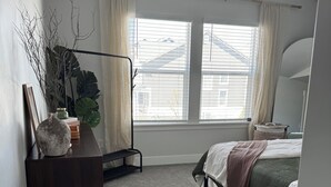 A bedroom with a bed, a bedside table, a dresser, a window with curtains, a wall clock, and a small plant. - The Windy Sage - peaceful and family friendly home for any event; sleeps 10+ (Eagle Mountain)