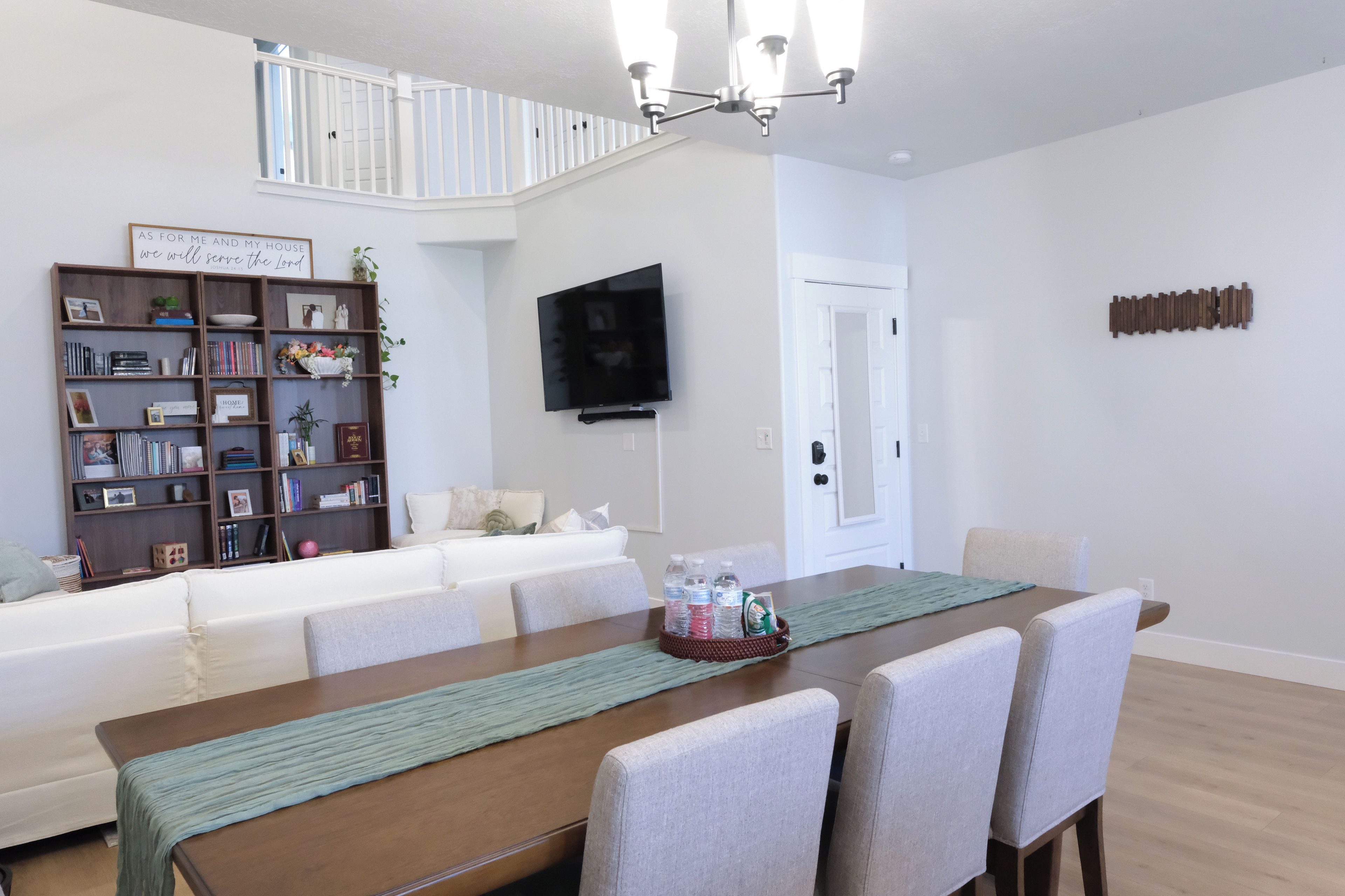 A dining area with a wooden table, chairs, a television mounted on the wall, and a bookshelf filled with books and decorative items.