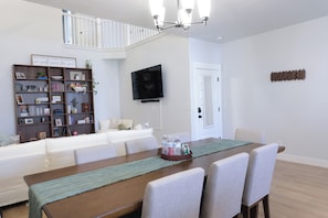Un comedor con una mesa de madera, sillas, un televisor montado en la pared y una estantería llena de libros y objetos decorativos.