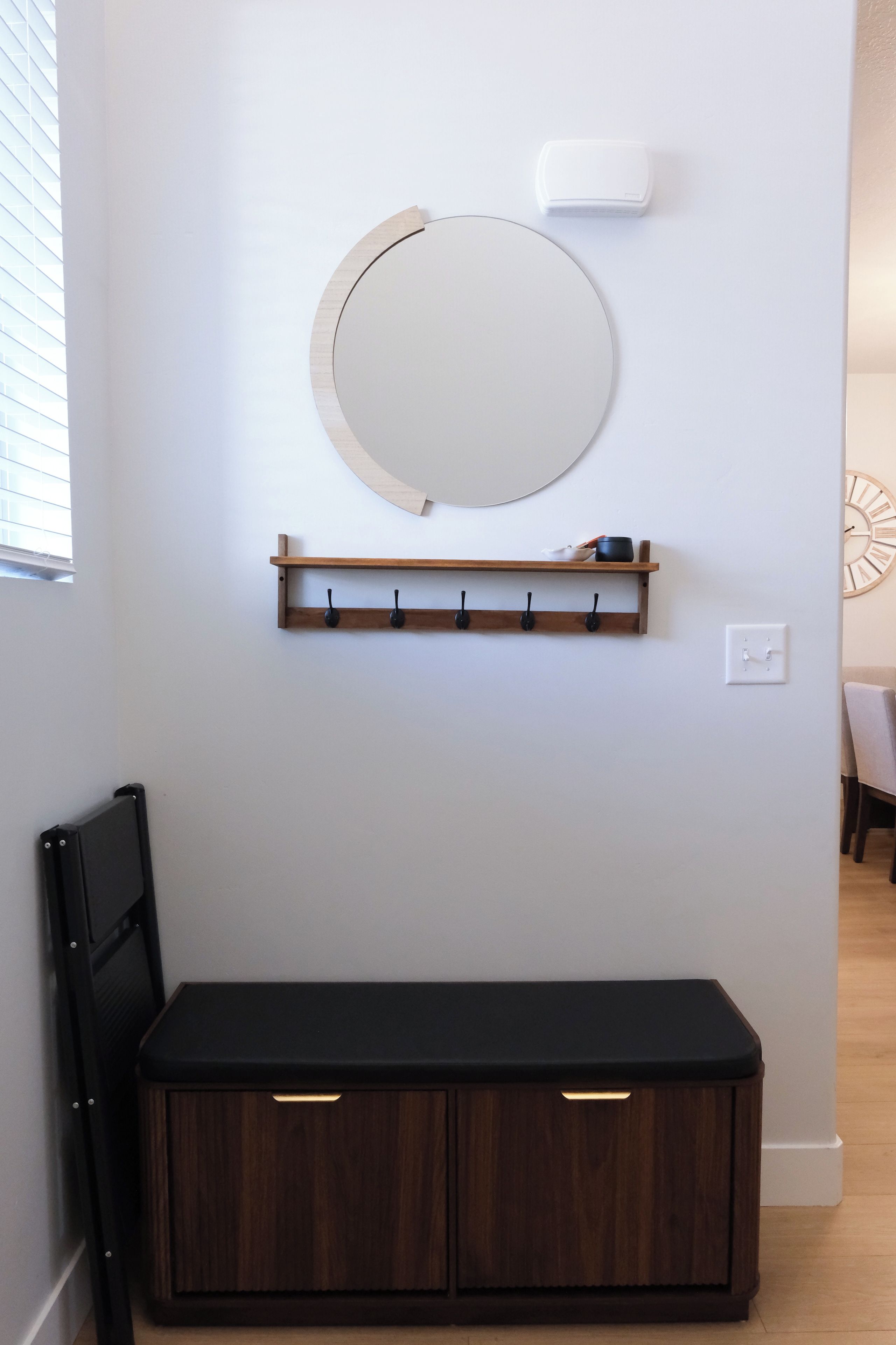 Quarto com um espelho circular fixado na parede, prateleira de madeira com ganchos e um banco de cor escura.