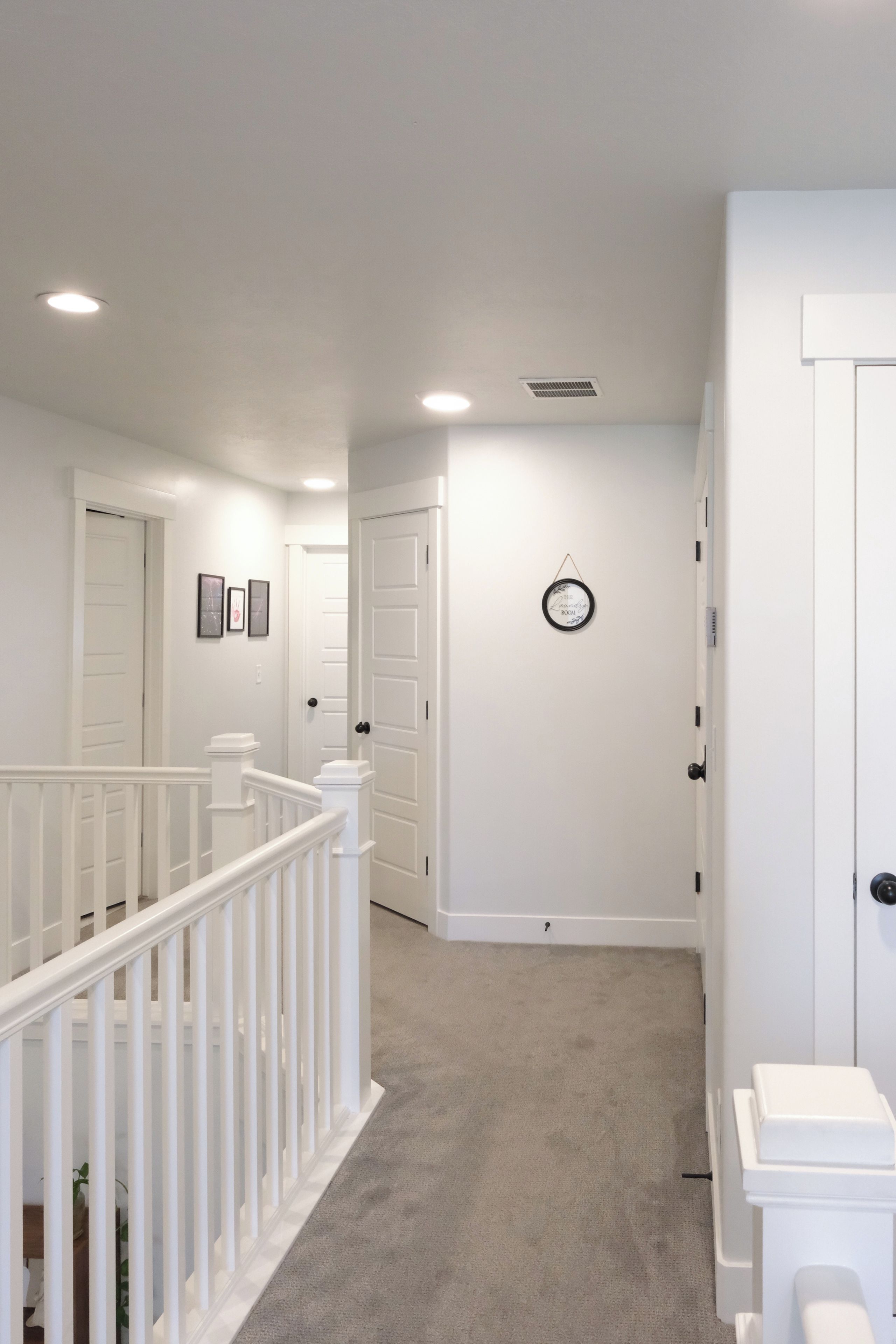 A hallway with white walls, a carpeted floor, and a white railing. There are doors on both sides, and a small round clock on the wall.