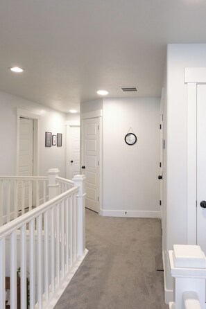 A hallway with white walls, a carpeted floor, and a white railing. There are doors on both sides, and a small round clock on the wall.