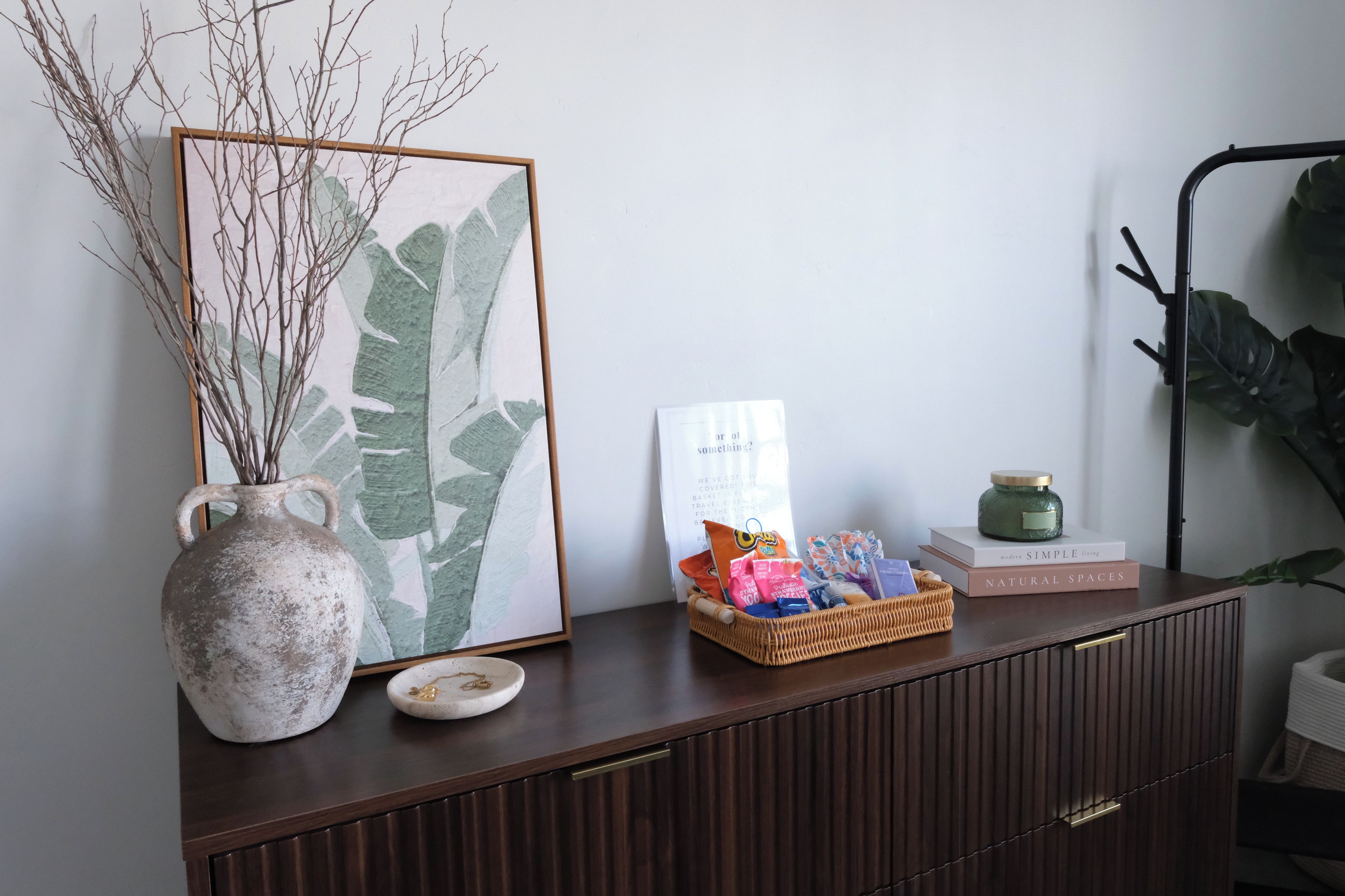 A wooden cabinet with a framed artwork, a vase with branches, a basket of snacks, a candle, and a book titled 'NATURAL SPACES'.
