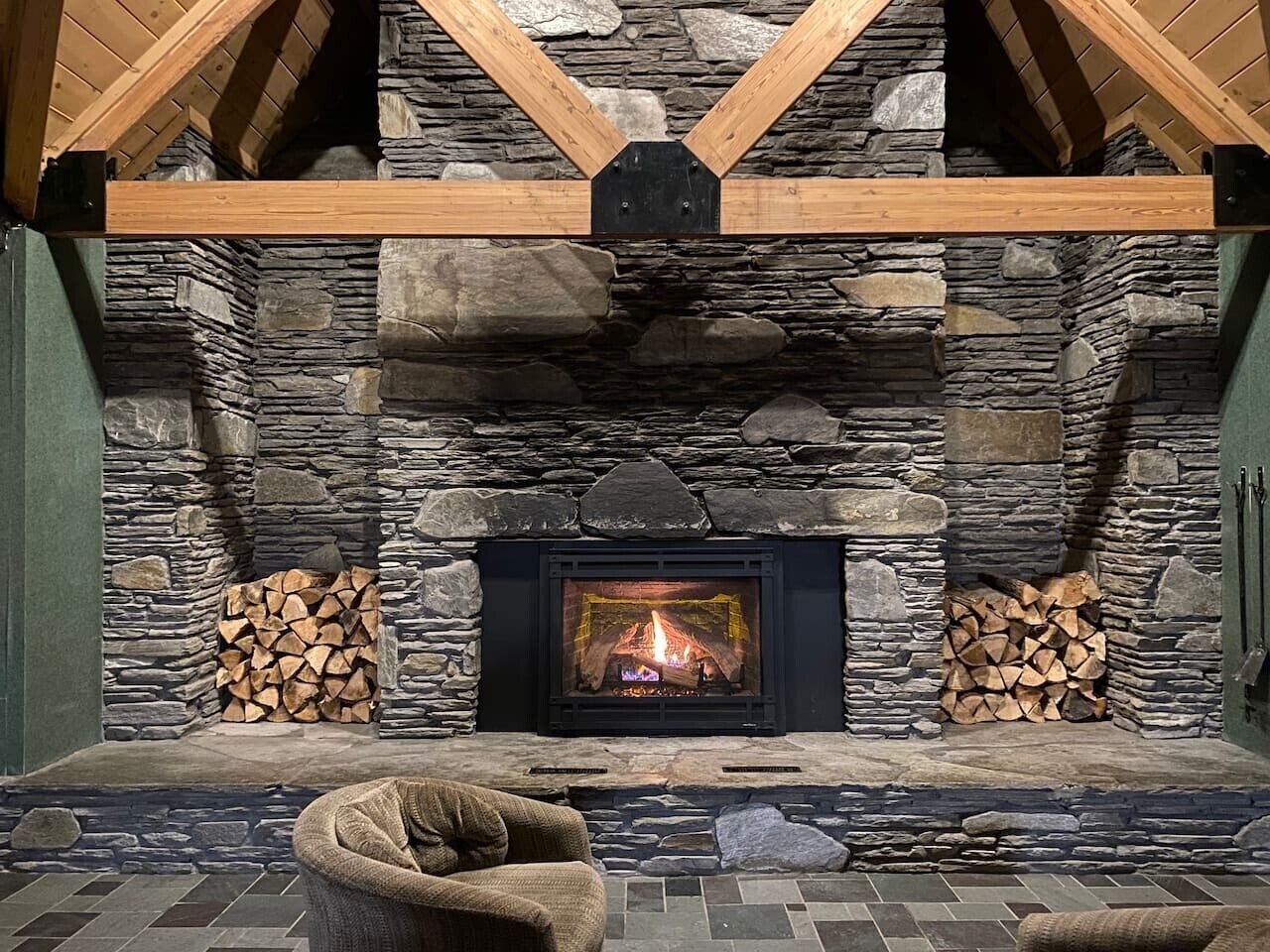 A stone fireplace with a fire, flanked by stacked firewood, in a room with a stone wall and wooden beams.