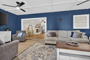A living room with a sofa, armchair, and a coffee table. There is a fireplace, a ceiling fan, and a television mounted on the wall. - Sandy Shores Pool & Spa Retreat w/Hot Tub/Beach (Summerton)