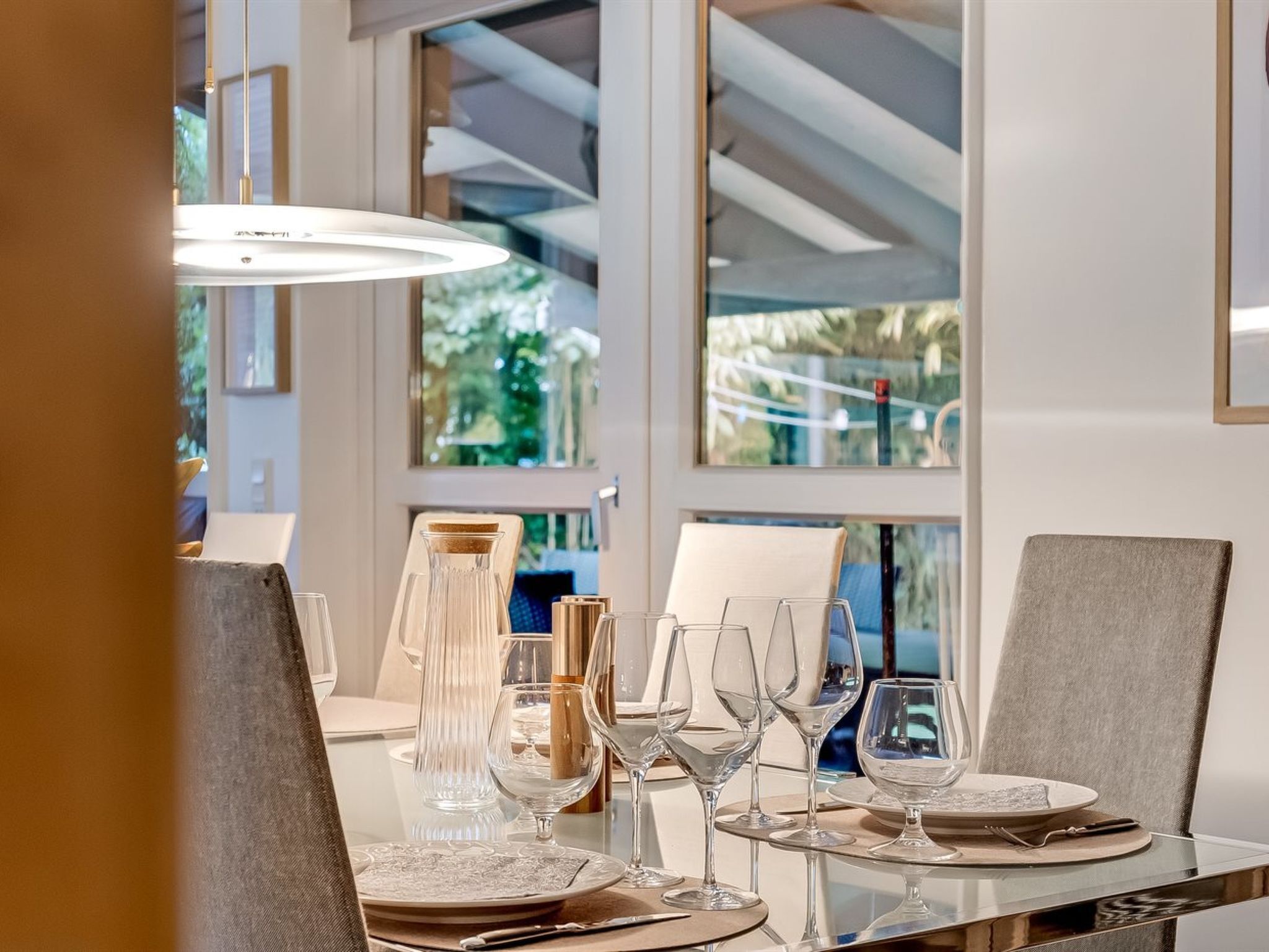 Une table à manger dressée avec de la vaisselle en verre transparent et des assiettes, un chandelier et une suspension moderne.