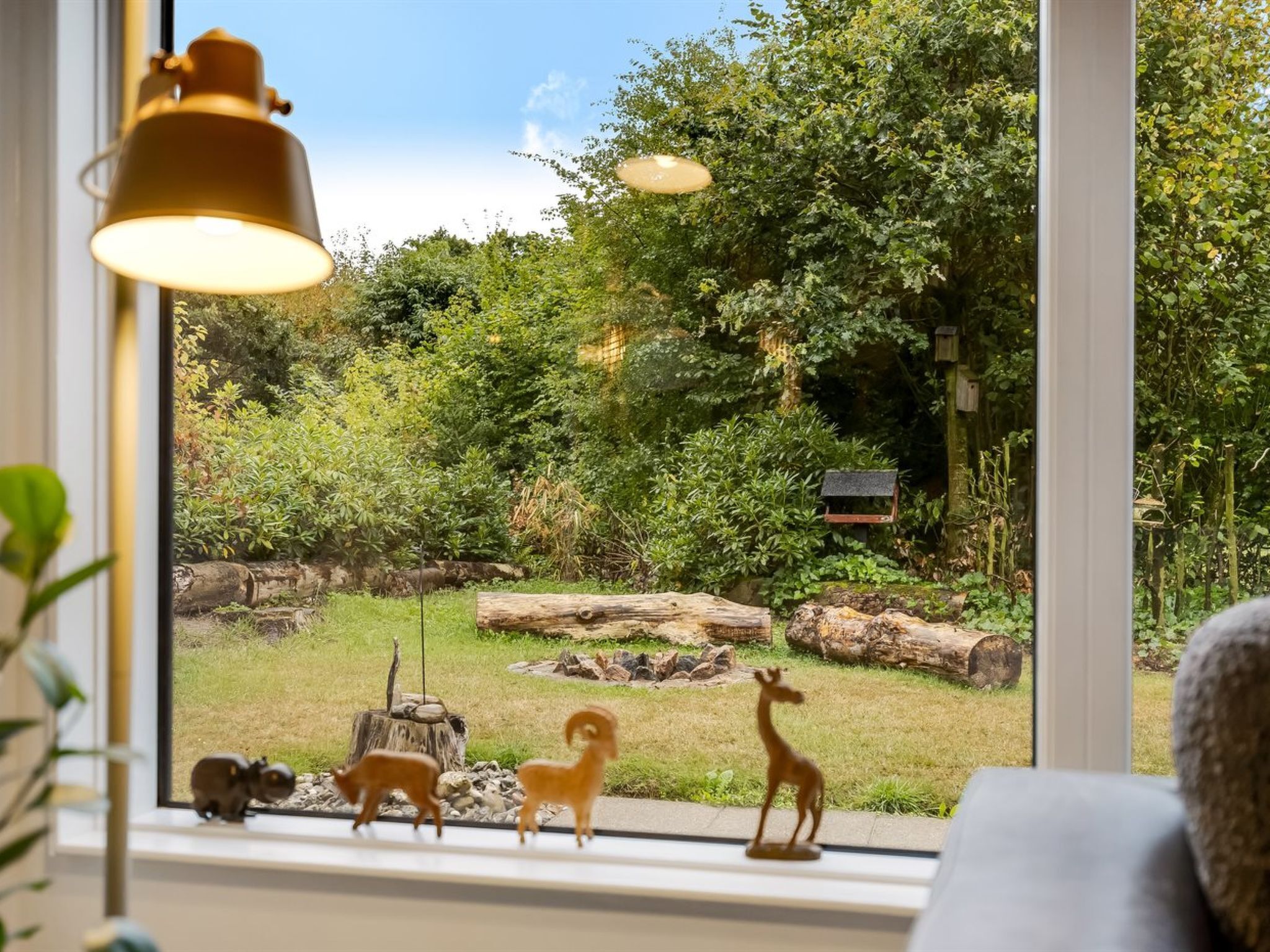 Een raam met uitzicht op een tuin, waar een hert, een vos en een vogelvoerder te zien zijn.