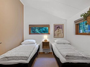 Quarto com duas camas de solteiro, uma pequena mesa de cabeceira com luminária, janela com vista para árvores e um quadro na parede.