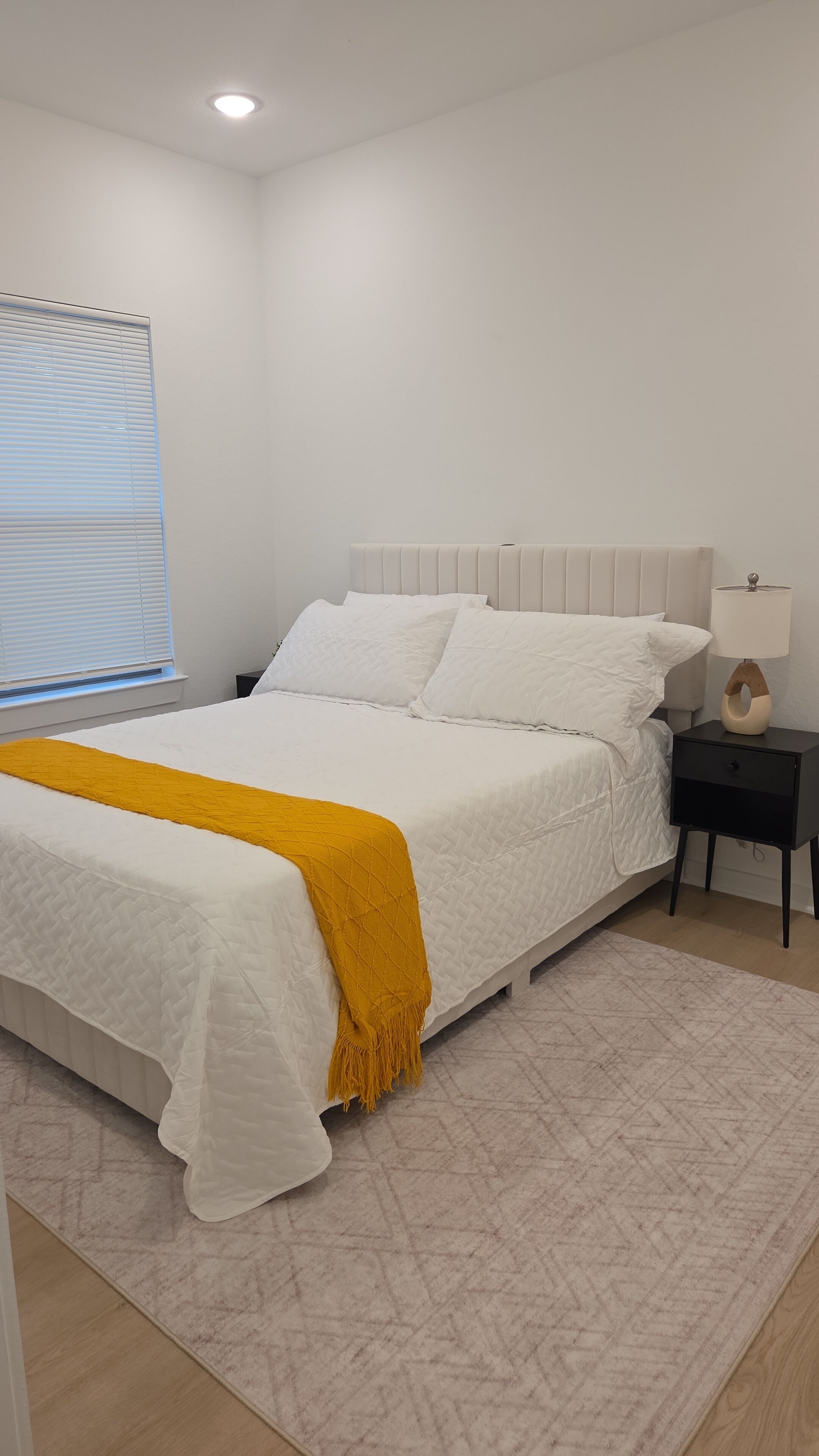 Quarto com cama, mesa de cabeceira, abajur e janela com persianas.