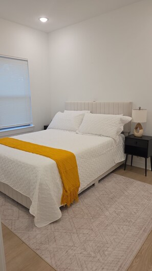 Quarto com cama, mesa de cabeceira, abajur e janela com persianas.