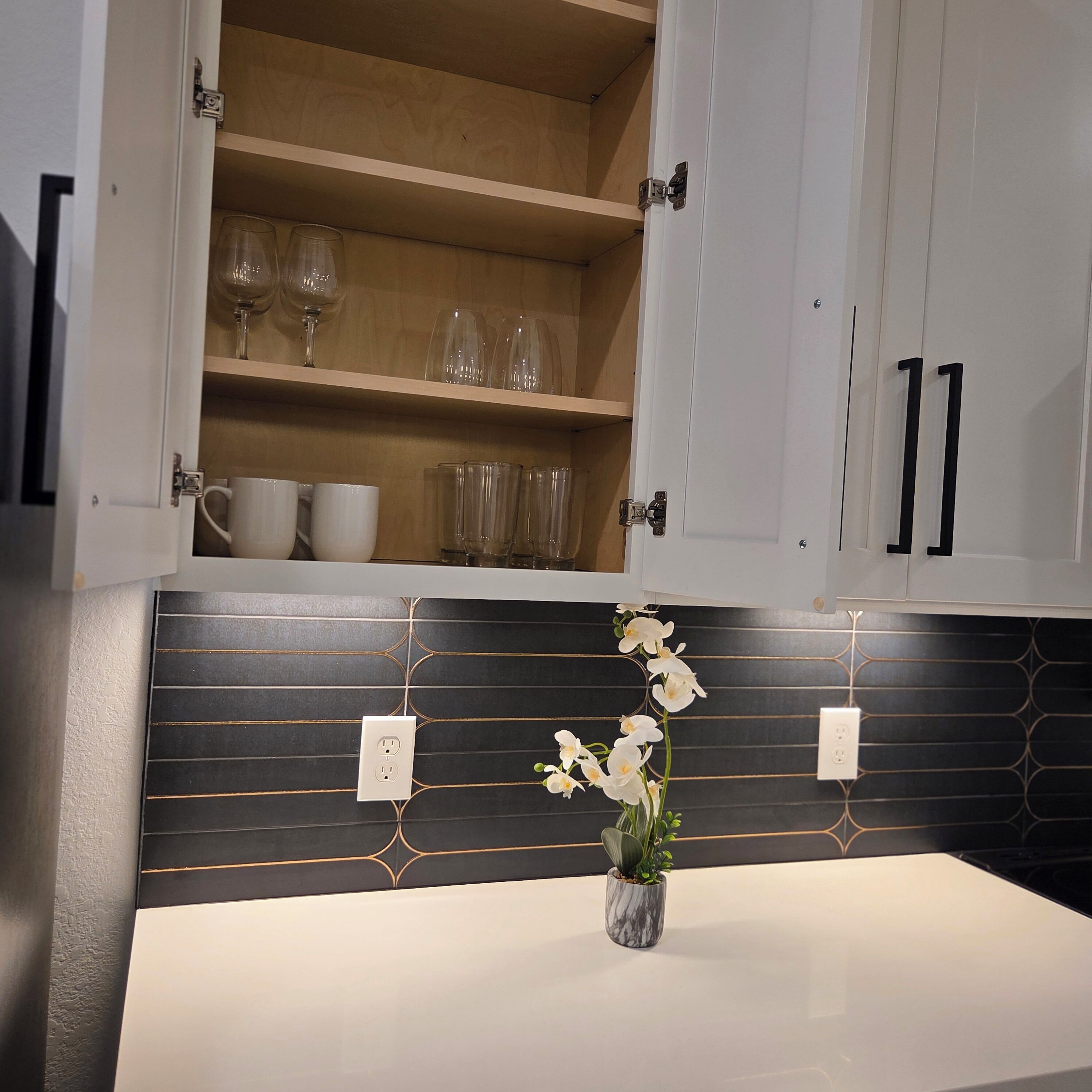 Et køkken med en hvid bordplade, en backsplash i sort og guld, samt et skab med vinglas og kopper.
