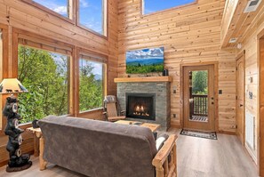 A log cabin interior with a fireplace, a sofa, a lamp, and a view of the outdoors. - Chestnut Tower – Incredible Mount LeConte Views, Hot Tub & Game Room (Gatlinburg)