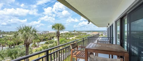 A balcony with a wooden table and chairs, a railing, and a view of a green landscape with palm trees.