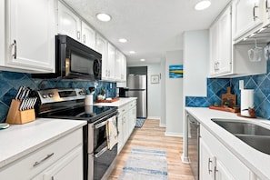 A modern kitchen with white cabinetry, a black microwave, stainless steel appliances, and a blue-tiled backsplash. - Ocean House 317: 2BR, 2BA beachfront condo with pool, balcony, views (St. Augustine)