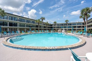A large outdoor swimming pool with lounge chairs and palm trees, surrounded by a multi-story hotel building. - Ocean House 317: 2BR, 2BA beachfront condo with pool, balcony, views (St. Augustine)