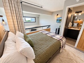 A bedroom with a bed, a window, a bedside table, and a bathroom visible through an open door.
