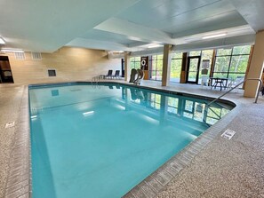An indoor swimming pool with clear water, surrounded by a tiled floor and walls, with windows providing natural light and views of the outdoors.