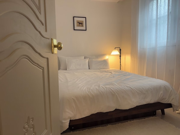 A bedroom with a bed, a door, a wall-mounted picture, and a bedside lamp.