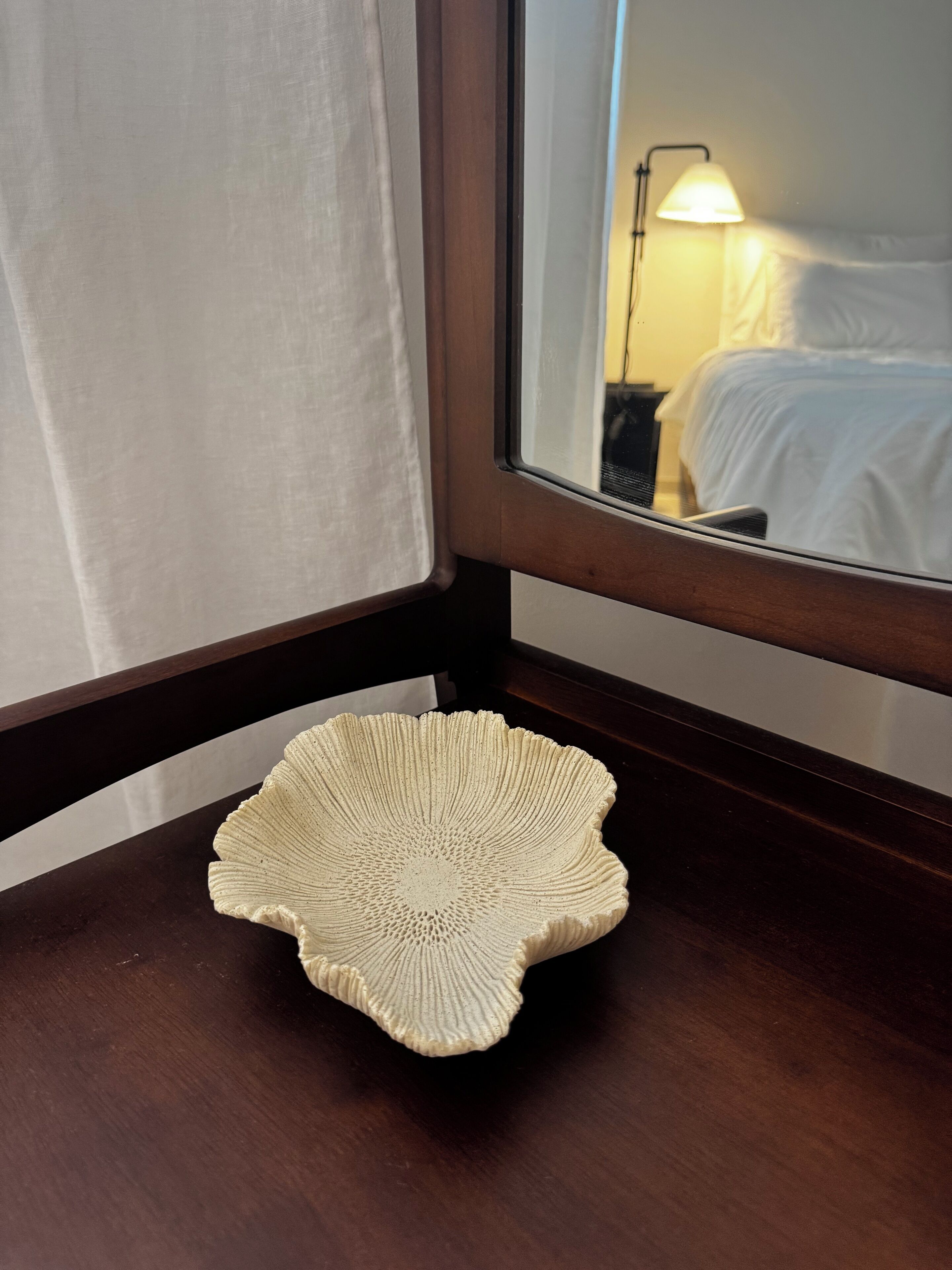 Een houten nachtkastje met een wit object met textuur, een spiegel die een bed en een lamp reflecteert, en een raam met witte gordijnen.
