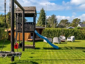 A playground with a wooden play structure, including a slide and swings, set in a grassy yard with white chairs and a hedge in the background. - "Rəzvan" -  from the sea by Interhome (Hadsund)