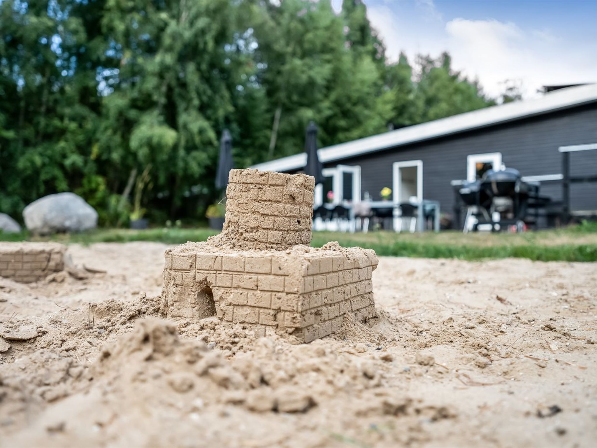 A sandcastle with a chimney, set against a backdrop of a building and trees.