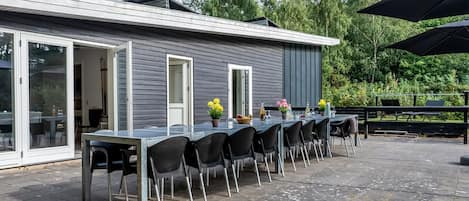 An outdoor patio with a long table and chairs, a glass door entrance, and a view of trees.