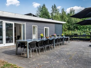 Eine Außenpatio mit einem langen Tisch und Stühlen, einem Glastür-Eingang und Blick auf Bäume.