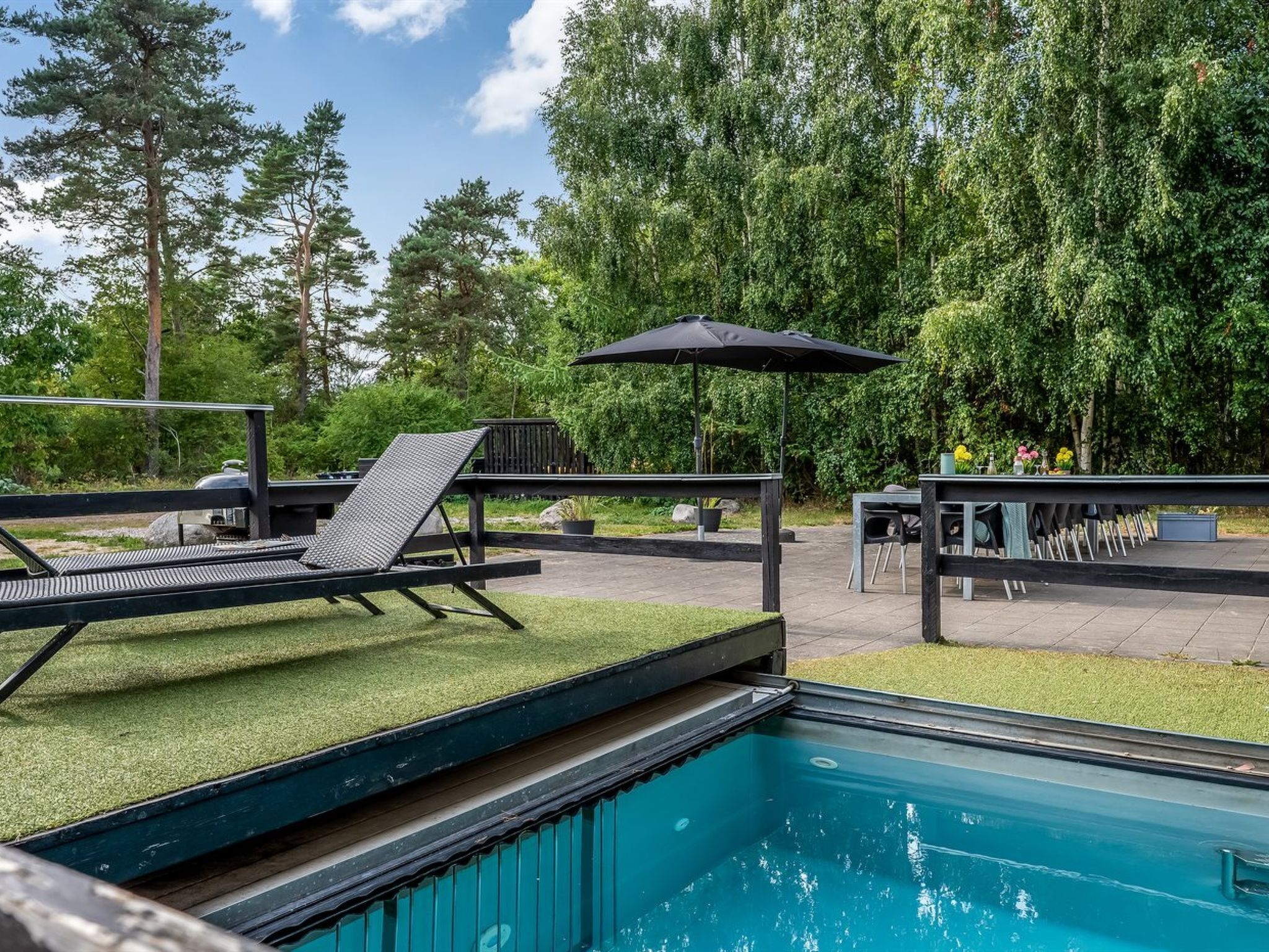 Une piscine entourée d’une terrasse et de nombreux arbres, avec des chaises longues et un parasol à proximité.