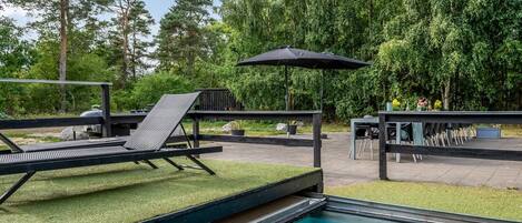 Une piscine entourée d’une terrasse et de nombreux arbres, avec des chaises longues et un parasol à proximité.