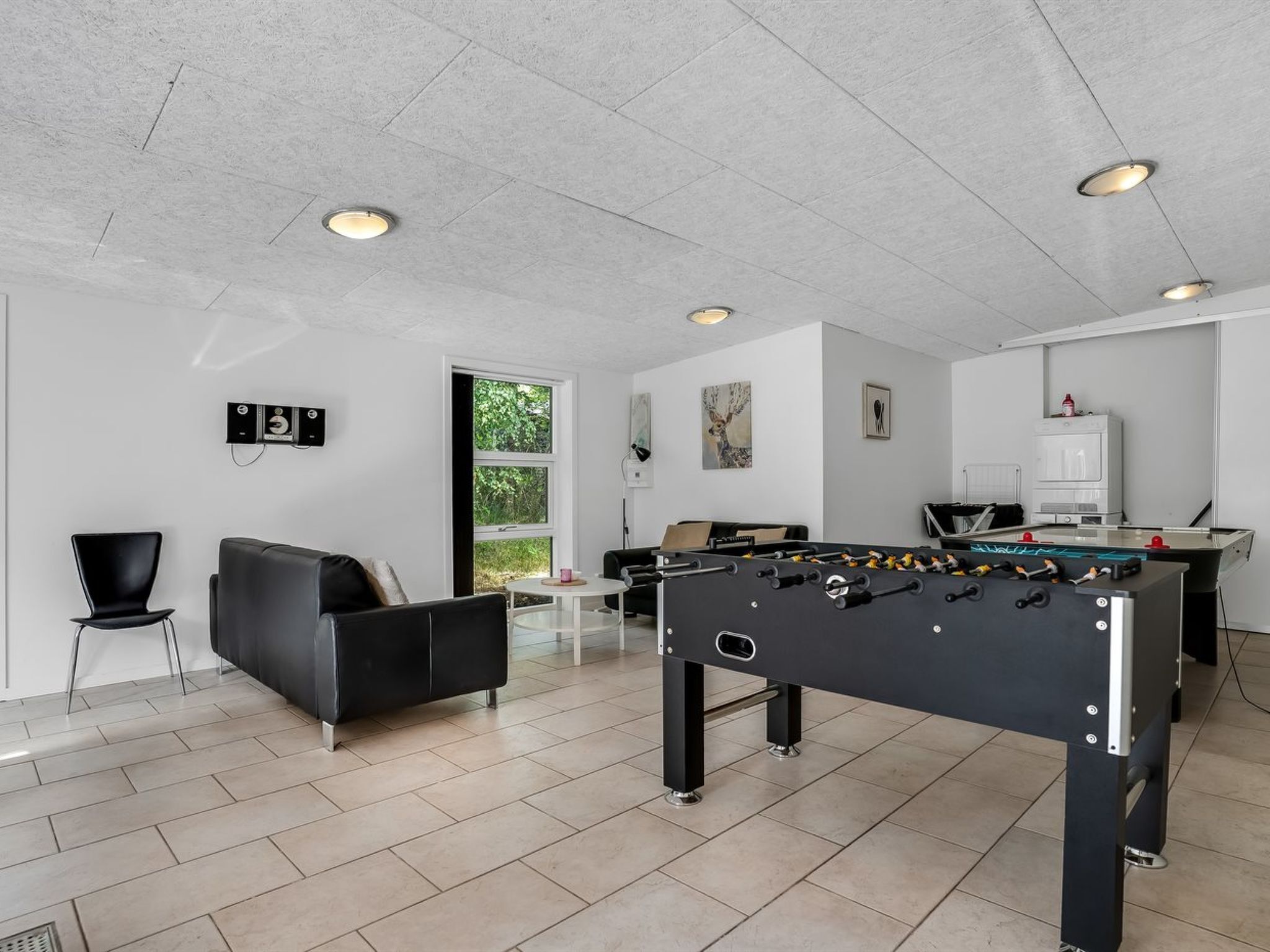 A modern living room with a foosball table, a sofa, a coffee table, and a window with a view of greenery.