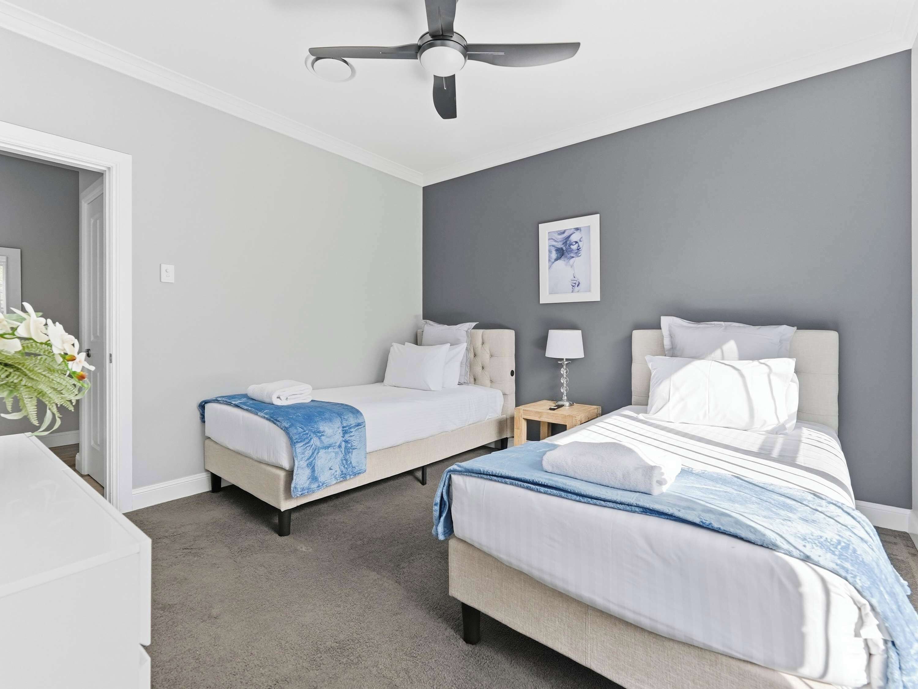 A hotel room with two beds, a ceiling fan, a bedside table with a lamp, and a framed artwork on the wall.