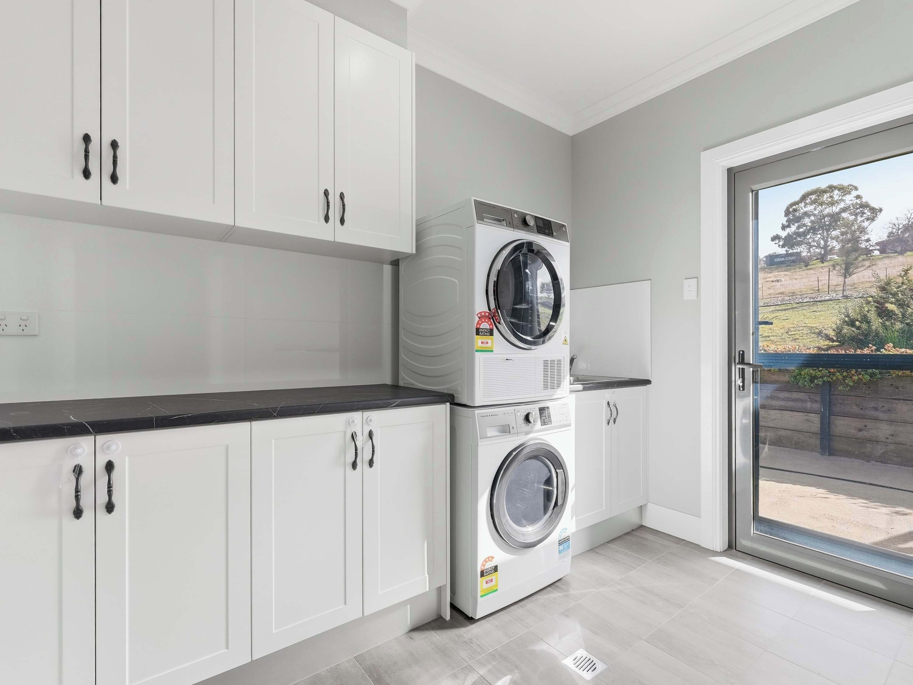 A laundry room with stacked washing machines and dryers, white cabinets, a black countertop, and a view through a glass door.