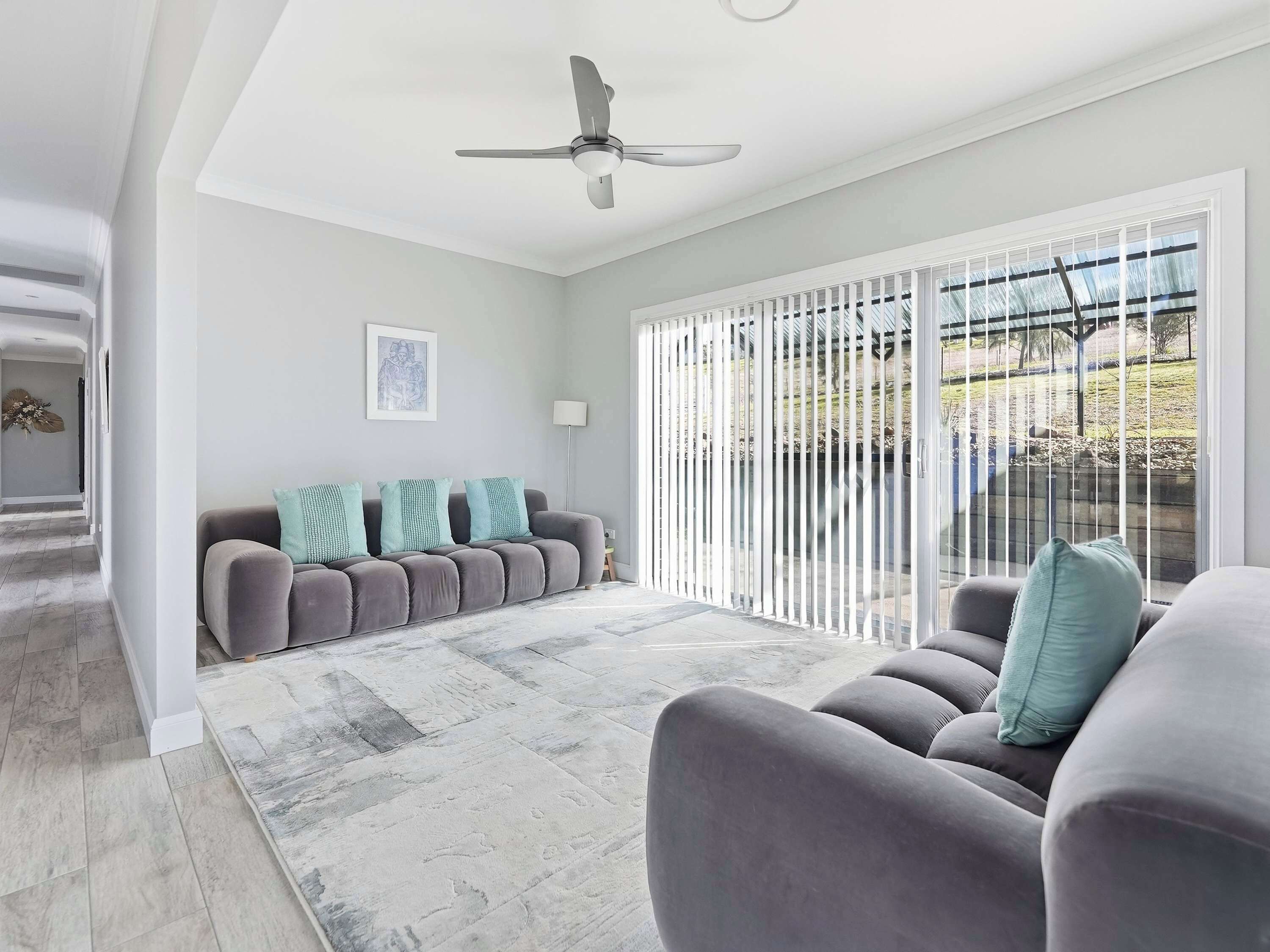 A modern living room with a grey sofa set, a ceiling fan, and a view through a sliding glass door to an outdoor area.