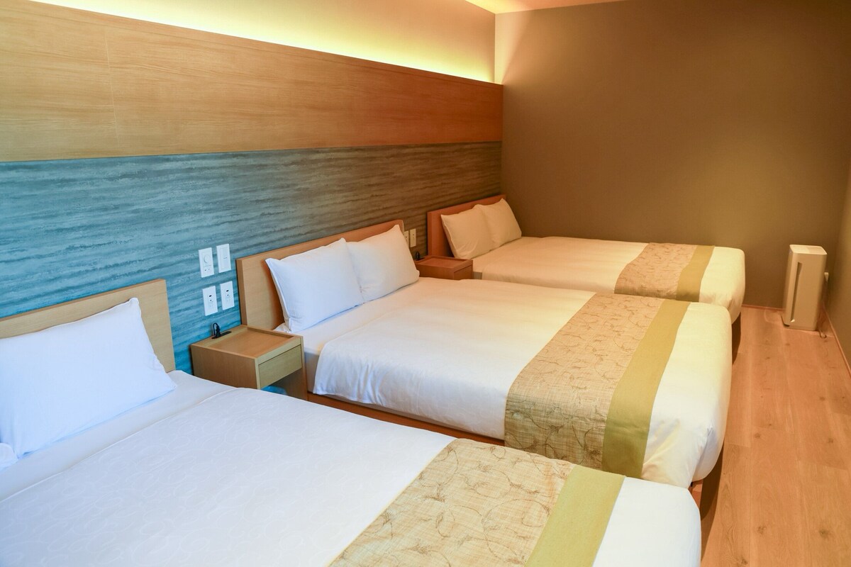 Habitación de hotel con tres camas, cabecera de madera, mesita de noche y pared con diseño texturizado.
