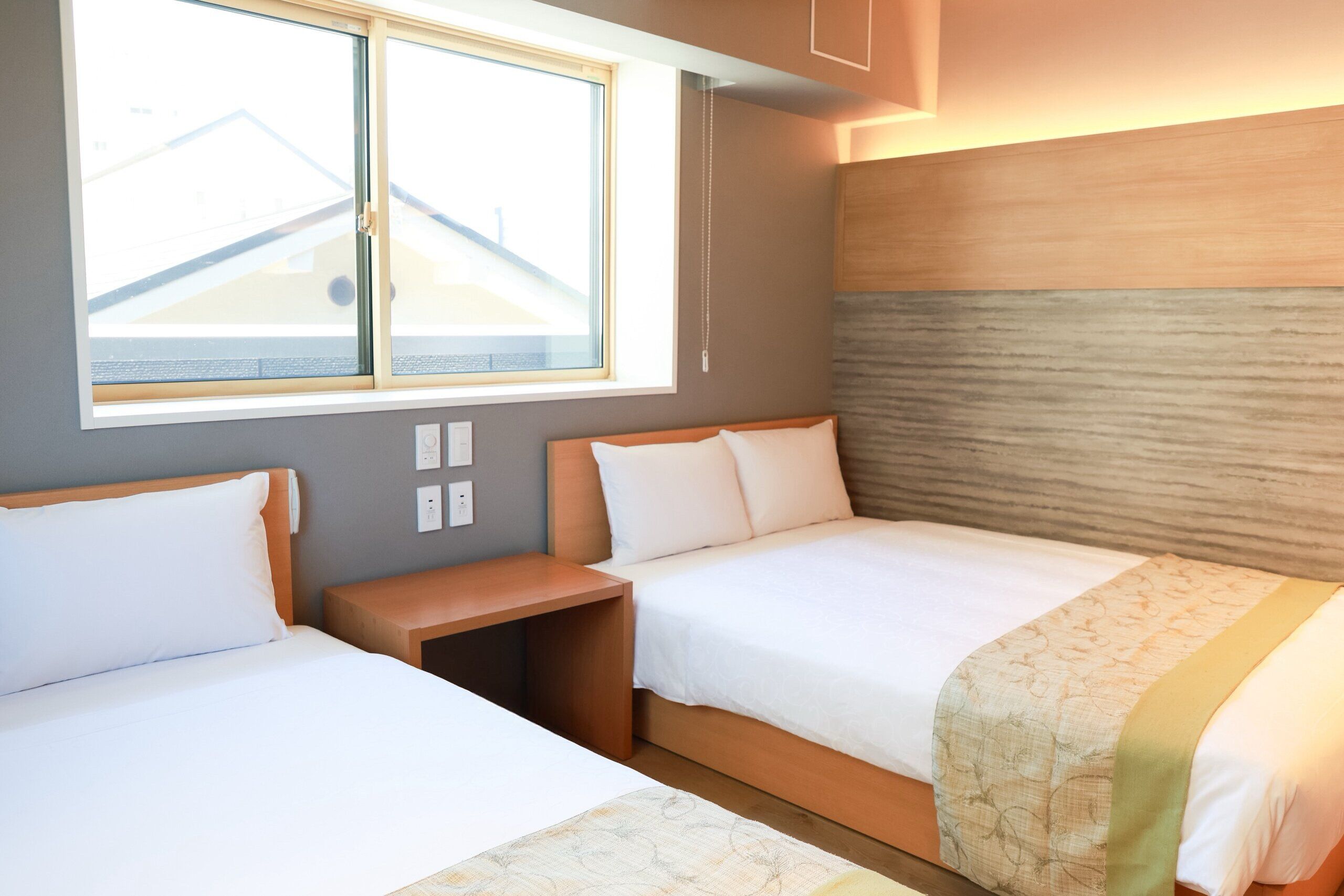 A hotel room with two beds, a nightstand, and a window with a view of buildings.