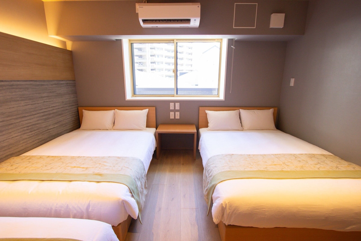 Habitación de hotel con dos camas individuales, una ventana que ofrece vistas a edificios y un equipo de aire acondicionado sobre la ventana.