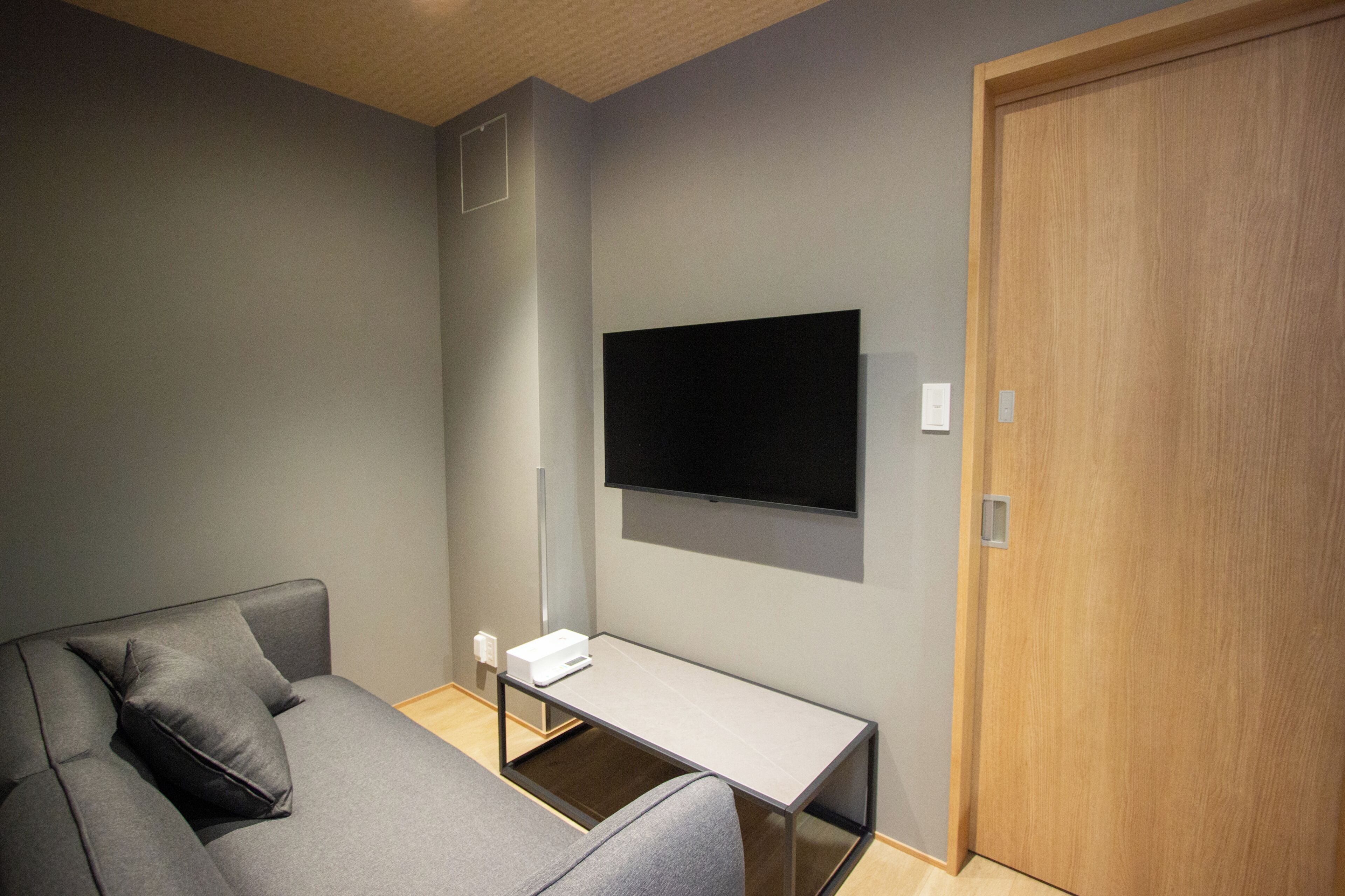 A modern room with a grey sofa, a small table, a flat-screen TV mounted on the wall, and a wooden door.