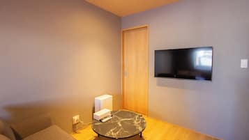 Ein Zimmer mit einem an der Wand befestigten Flachbildfernseher, einem kleinen runden Tisch mit einer Fernbedienung und einer Couch.