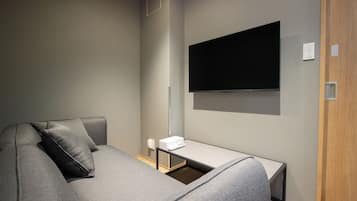 Une chambre d’hôtel moderne avec un canapé gris, un téléviseur à écran plat fixé au mur et une petite table sur laquelle se trouve une télécommande.