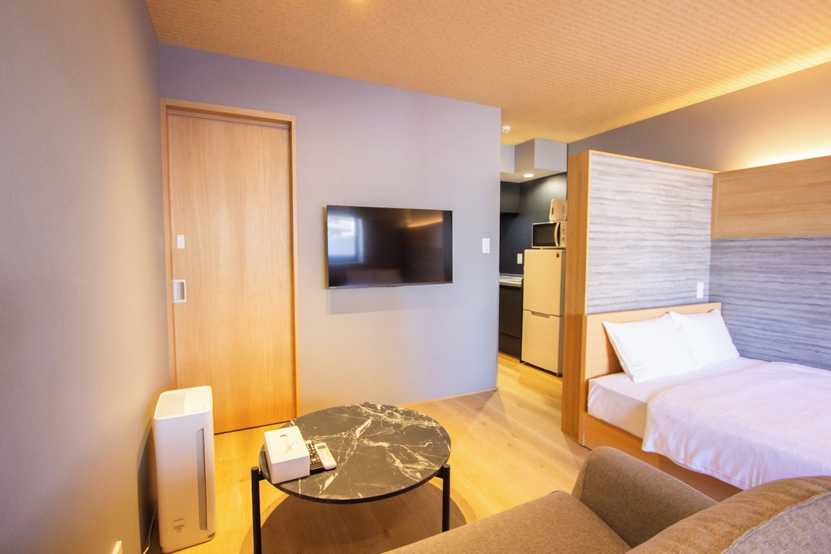 Habitación de hotel con cama, televisión, escritorio, silla y una kitchenette al fondo.