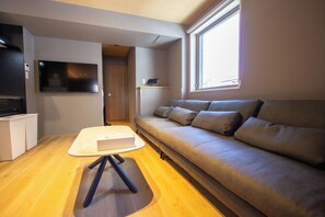A compact living space with a sofa, a flat-screen TV, a small table, and a kitchenette area. - Nonette Hakata Haruyoshi (Fukuoka City)