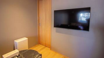 Una habitación con una puerta de madera cerrada, un televisor de pantalla plana montado en la pared, un dispositivo blanco en el suelo y una mesita redonda con un control remoto encima.